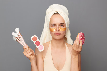 Sad upset woman wrapped in towel standing cosmetics brushes and palette of eye shadows and tasty donut, expressing sorrow, can't eat, being on diet, posing isolated over gray background.の写真素材