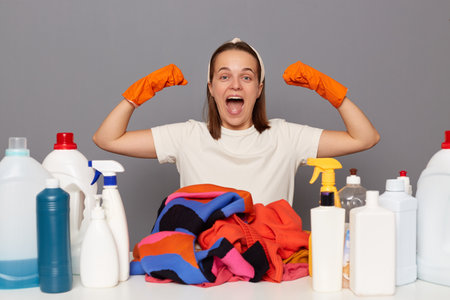 Excited overjoyed young adult woman wears headband, orange gloves and casual t shirt, sitting at table among different detergents, raised arms, showing her power, isolated over gray background.の写真素材
