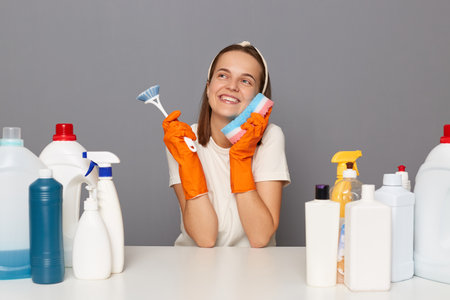 Indoor shot of smiling positive happy Caucasian woman wears headband, gloves and casual t shirt, posing isolated over gray background, smiles broadly, dreaming, surrounded with bottles of detergents.の写真素材