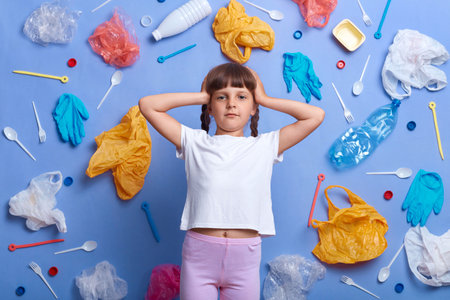 Worried upset little girl with braids wearing white t shirt posing against much plastic wastes on blue wall, standing hand on head, looking at camera, doesn't want listen about polluting environment.の写真素材
