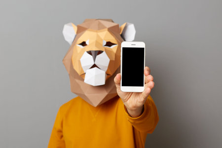 Portrait of man with lion mask with wearing casual style orange jumper posing isolated over gray background, showing cell phone with empty black display, copy space for promotion.の写真素材