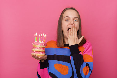 Photo of excited amazed woman with brown hair wearing stylish jumper, covering mouth with palm, having surprise on her birthday party, holding donut with candles isolated over pink background.の写真素材