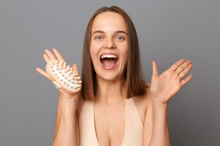 Indoor shot of excited joyful woman uses dry body brush wants to have smooth clean skin, yelling with excitement, poses against gray background, posing with open mouth.の写真素材