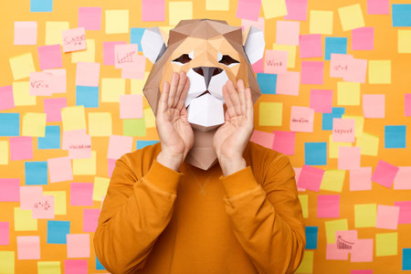 Horizontal shot of man wearing orange jumper and paper lion mask posing against yellow wall with stickers, keeps hands near face, screaming, making announcement.の写真素材