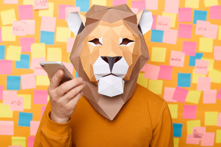 Image of man wearing orange sweater and paper lion mask posing isolated over yellow background, holding smart phone in hands, using mobile phone, recording voice message.の写真素材