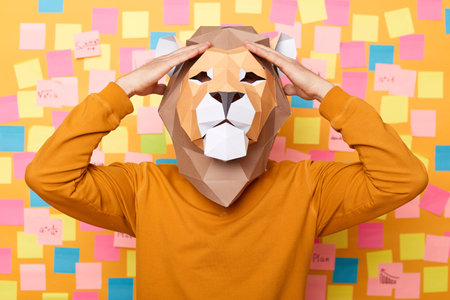 Indoor shot of sick unhealthy man wearing orange sweater and paper lion mask posing isolated over yellow background, standing holding hands on head, feels unwell.の写真素材