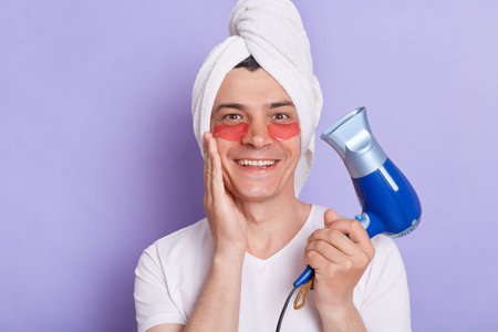 Funny Caucasian man wrapped towel on head doing morning cosmetology procedures, posing with patches under eyes isolated over purple background, holding hairdryer with positive expression.の写真素材