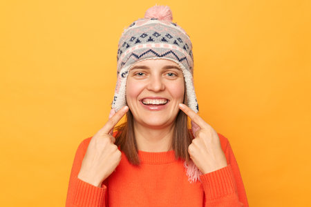Horizontal shot of delighted young adult woman wearing funny earflap hat and orange casual jumper...