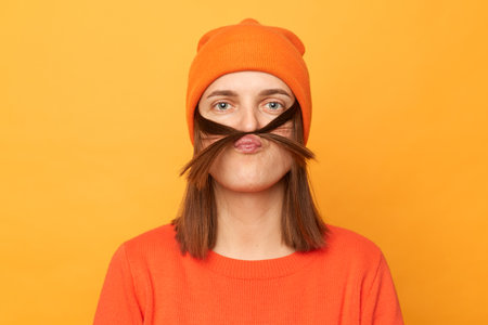 Portrait of funny positive hipster woman wearing orange sweater and hat posing isolated over yellow...