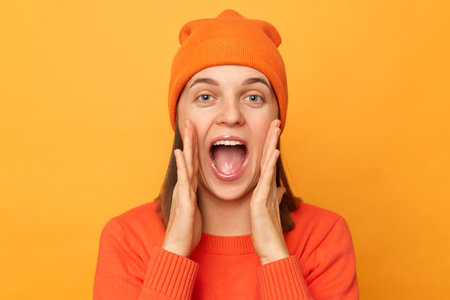 Indoor shot of amazed positive young adult hipster woman wearing orange sweater and hat posing...