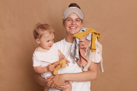 Photo of cheerful woman wearing white T- shirt and blindfold holding her baby daughter in hands isolated over brown background, expressing happiness, enjoying maternity leave.の写真素材