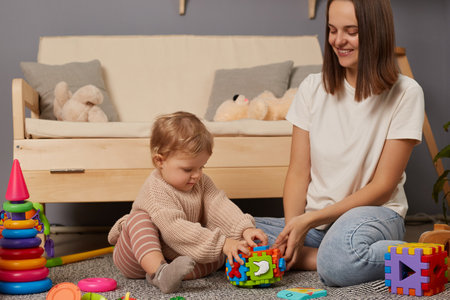 Photo of woman with dark hair sitting on floor, looking at her playing baby with smile, toddler girl studying new interesting toy, playing with colorful sorter.の写真素材
