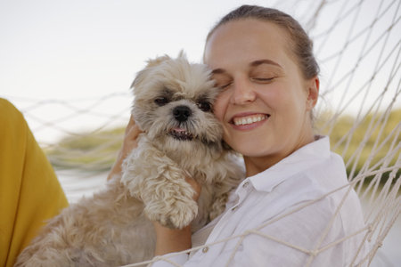 Smiling delighted satisfied Caucasian woman wearing white shirt lies in a hammock with Pekingese dog on the bank of the river, female keeps eyes closed, hugging her pet, enjoying weekend in nature.の写真素材