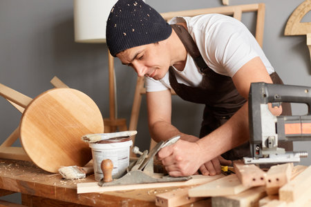 Image of handsome guy carpenter wearing apron and cap working with plane on wooden workplace...