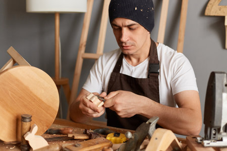 Portrait of carpenter man in brown apron making a handmade wooden toy in a home workshop, handyman carves a toy in wood with a special tool, joiner in the workshop.の写真素材
