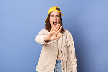 Portrait of annoyed teenager girl wearing baseball cap and jacket posing isolated over blue background, expressing negative emotions, showing stop gesture, screaming.の写真素材