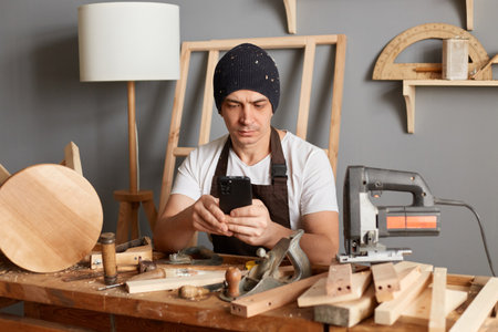 Image of handsome Caucasian man carpenter wearing white t-shirt, black cap and brown apron, sitting at table and using smart phone, finds new creative ideas for his joinery work.の写真素材