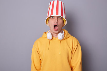 Portrait of amazed shocked emotional hipster man wears yellow hoodie standing against gray wall...