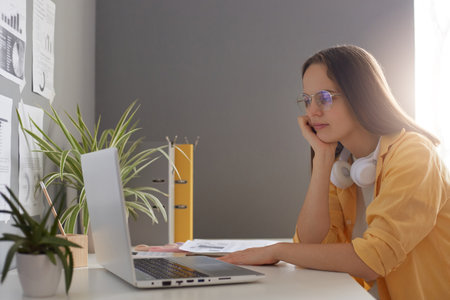 Horizontal shot of calm beautiful woman dressed in yellow shirt using laptop computer and sitting at...