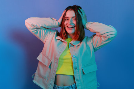 Indoor shot of smiling attractive, young girl wearing casual jacket standing isolated over blue neon light background, looking at camera, keeps hand on her head.の写真素材