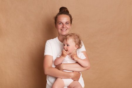 Portrait of delighted smiling woman with bun hairstyle wearing white T- shirt standing with her baby...
