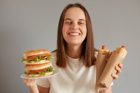 Portrait of happy smiling woman holding hot dog and burger wearing white T-shirt posing isolated over gray background, looking at camera with toothy smile, enjoying eating fast food.の写真素材