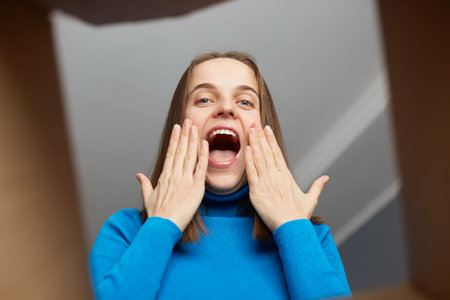Beautiful excited woman customer wearing blue turtleneck opening parcel, looking inside package with amazed look, touching her face in excitement, receiving online order, bottom view.の写真素材