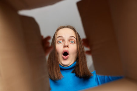 Shocked amazed Caucasian woman customer with brown hair wearing blue turtleneck opening parcel, looking inside cardboard box with shocked face, saying wow, unexpected package.の写真素材
