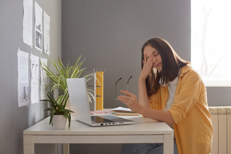 Tired female freelancer sitting in office in front of laptop, taking off her glasses, feels eyes...