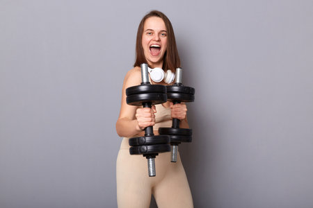 Horizontal shot of happy crazy funny woman wearing sportswear working out triceps and biceps with weights, showing dumbbells, screaming, has work out for her arms, posing isolated on gray backgroundの写真素材