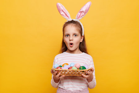 Portrait of amazed little girl wearing bunny ears holding Easter eggs in wicker basket isolated on...