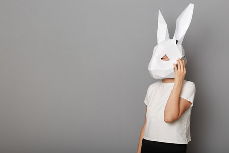 Image of unrecognizable woman wearing white t shirt and paper rabbit mask standing isolated over gray background, talking on smart phone, copy space for advertisement, mockup.の写真素材