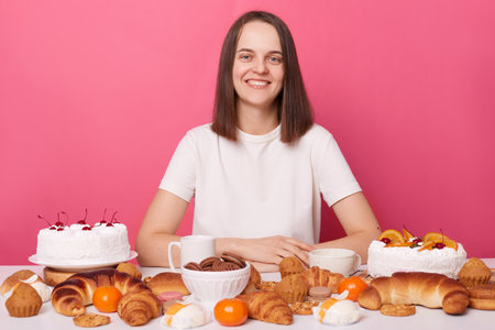 Portrait of lovely young woman with brown hair sitting at big table full of cookies, pastries and cakes, eats different desserts during holidays, posing isolated over pink background.の写真素材