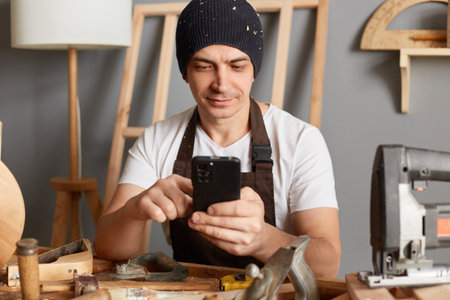 Indoor shot of handsome caucasian young adult man in cap and apron using his mobile phone in workshop, having break, chatting with friends, checking social networks.の写真素材