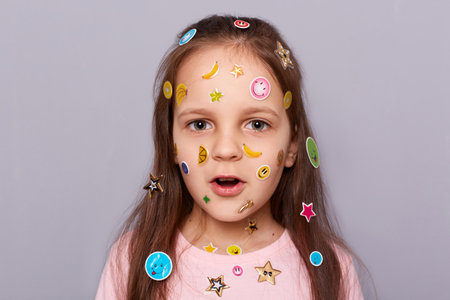 Closeup shot of shocked amazed little brown haired little girl covered with stickers looking at...