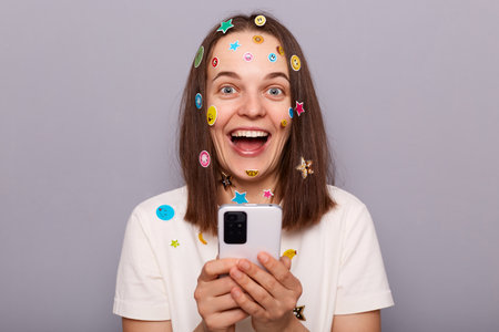 Indoor shot of surprised cheerful woman covered with funny stickers holding smartphone, winning online lottery, looking at camera with big joyful eyes, posing isolated over gray backgroundの写真素材
