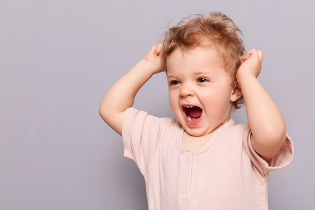 Portrait of laughing playful expressive cute blonde laughing infant girl with wavy hair screaming while playing, being in good mood, looking away, standing isolated over gray backgrond, copy space.の写真素材