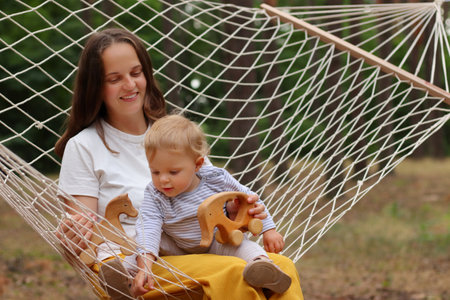 Portrait of happy smiling mother and child relaxing in hammock in forest, mom and daughter playing wooden eco toys, spending summer day, enjoying nature and fresh air.の写真素材