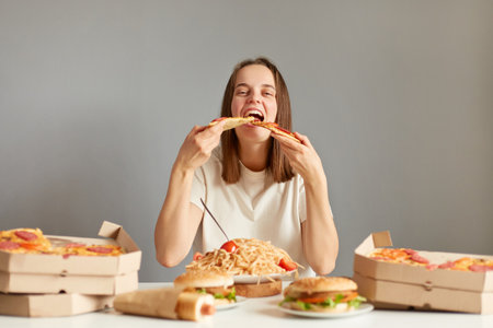 Portrait of extremely hungry woman with brown hair wearing white T-shirt sitting at table isolated over gray background, biting two slices of pizza same time.の写真素材