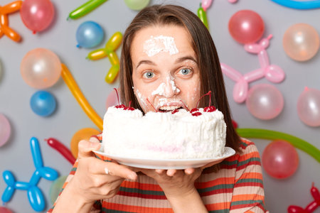 Positive happy beautiful woman in striped dress standing against gray wall decorated with colorful balloons biting her birthday cake after dipping face in cream.の写真素材