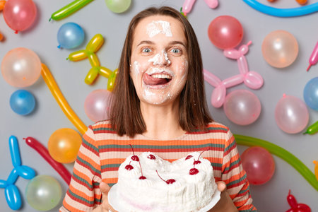 Attractive hungry birthday woman in striped dress standing against gray wall decorated with colorful...