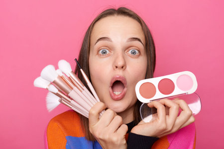 Shocked surprised woman wearing sweater holding brushes and powder posing isolated on pink background looking with big eyes, being amazed with new cosmetic products.の写真素材