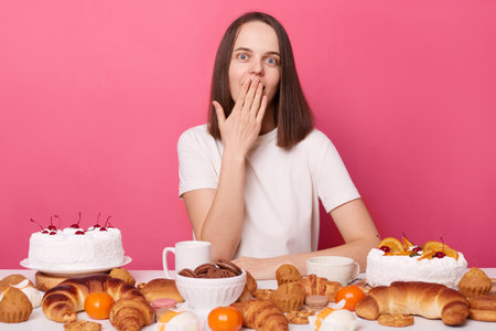 Puzzled surprised brown haired woman in white t shirt sitting at table with lots of desserts breaking diet covering mouth with palm isolated over pink background.の写真素材