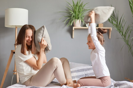 Funny pillow fight. Smiling happy mother and her little daughter playing in bed waking up in good playful mood rejoicing having fun enjoying weekend morning.の写真素材