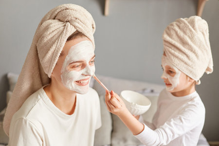 Indoor shot of smiling woman doing beauty procedures with her daughter, little kid applying mask to mommy face enjoying moment spending mother's day in spa.の写真素材