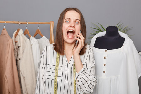 Shocked despair caucasian woman tailor talking on phone in sewing studio, standing near hangers with...