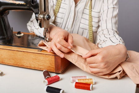 Unknown woman seamstress sewing clothing on her machine in tailor workshop, holding textile,...