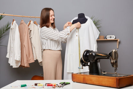 Satisfied professional seamstress measuring dress collar with tape during sewing process in atelier, enjoying working process, making handmade outfit in dressmaking workshop.の写真素材