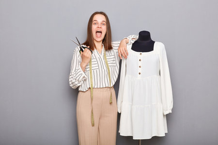 Excited caucasian female sewer standing near mannequin with new dress isolated over gray background...