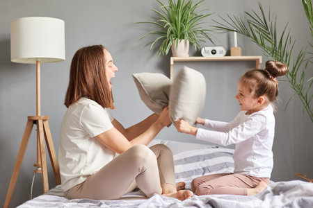 Excited amazed family mother and daughter having fun together playing fighting with pillows laughing sitting on bedroom on bed expressing happiness.の写真素材